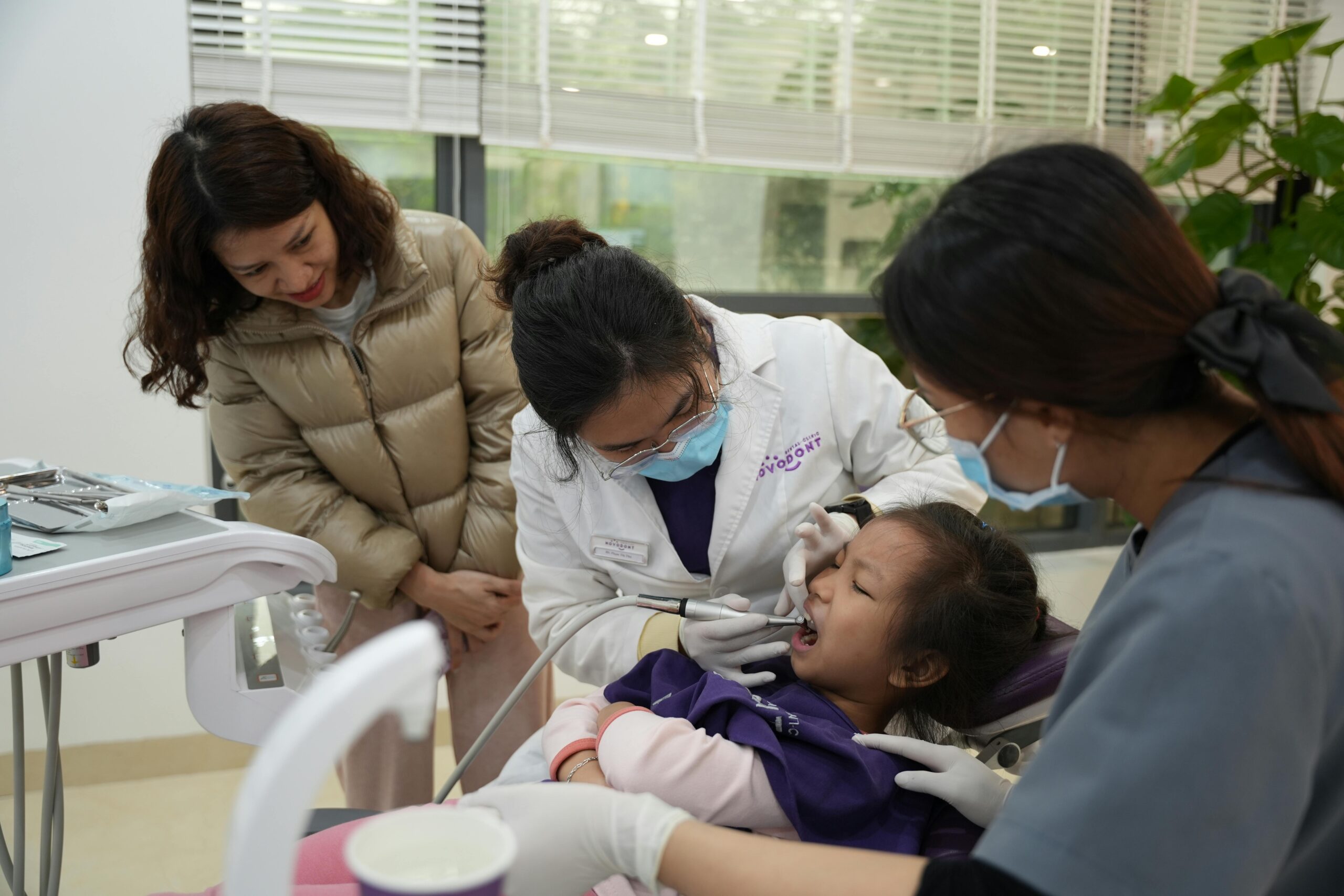 A dentist examines a young girl's teeth, with a parent and assistant observing in a modern clinic.